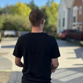 A man viewed from behind wearing a black short-sleeve t-shirt and light-colored sunglasses, standing on a paved outdoor walkway with blurred houses and trees in the background.