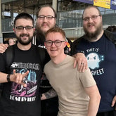 Four men posing together outdoors in front of a modern glass building. Three men are wearing graphic t-shirts: one in a black band shirt with 'ROSEVILLE' visible, one in a black shirt with a skull graphic