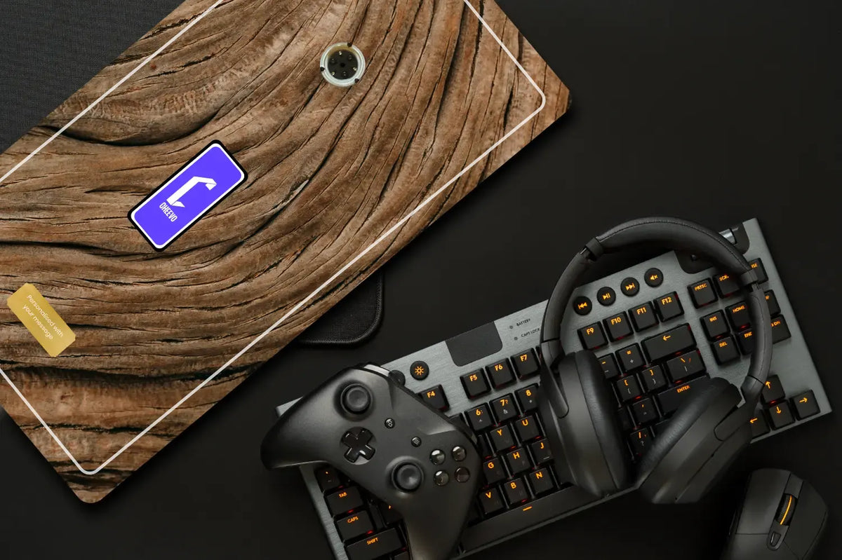 A top-down flat lay of a gaming setup on a dark surface, featuring a black mechanical keyboard with orange backlighting, a black wireless controller, over-ear headphones, and a mouse. A large rectangular wooden board with a printed circuit board design and a small gold rectangular piece lies diagonally in the upper left.
