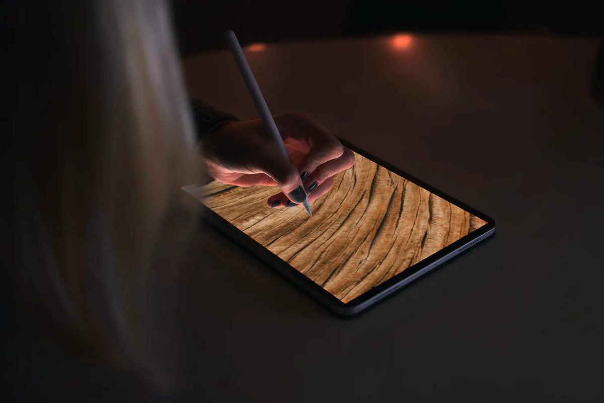 A person holds a stylus over a tablet displaying a wood grain texture, set against a dark, dimly lit background.