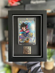 A framed display of Dragon Ball Super trading card, featuring the card inside a case and a QR code mounted in a black shadow box with a white border. The frame is being held up against a gaming setup, with shelves displaying collectibles and gaming accessories in the background.