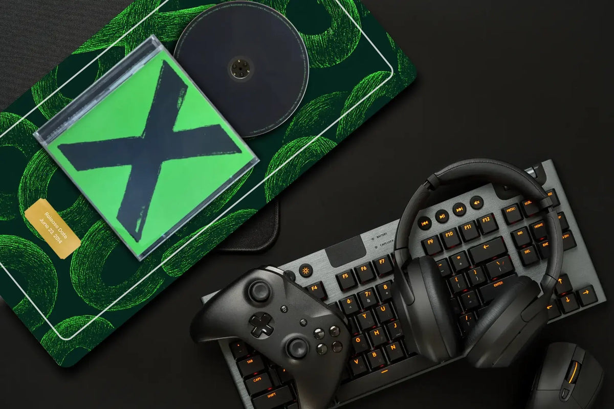 A flat-lay of gaming accessories on a dark surface, including a green patterned mouse pad with a clear CD case, a mechanical keyboard with orange-backlit keys, over-ear headphones, and a game controller.