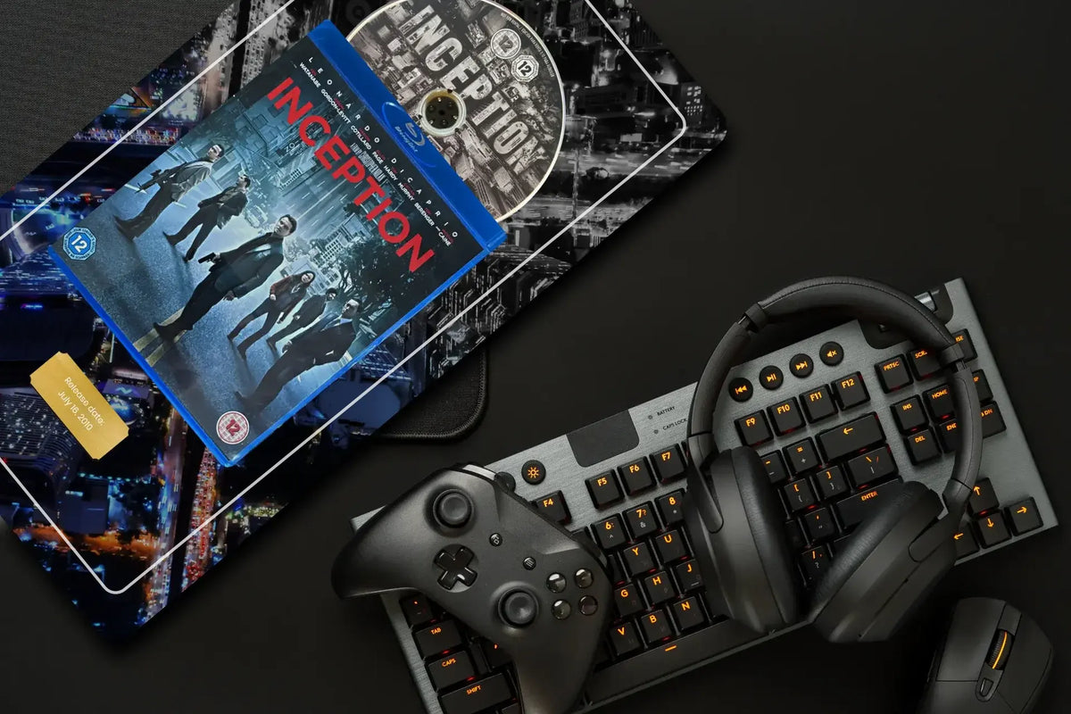 A flat-lay arrangement on a dark surface featuring a printed artwork with an Inception cityscape design, a blue Blu-ray case, a DVD, a black gaming keyboard with orange backlighting, over-ear headphones, and a black game controller.