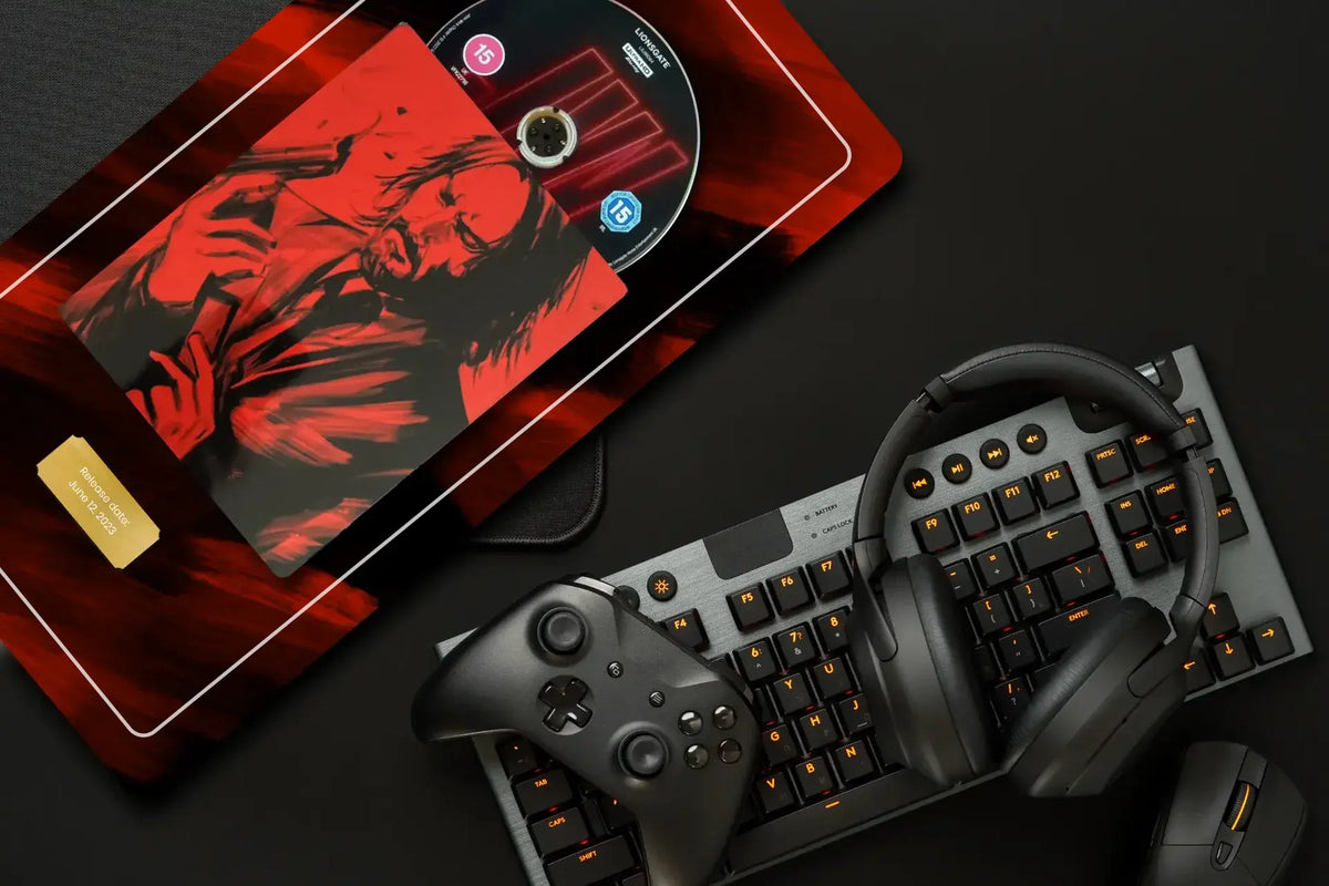 A styled flat-lay of gaming accessories on a dark surface, including a black keyboard with orange backlighting, over-ear headphones, a game controller, a mouse, and a red-and-black rectangular case or tray holding a printed card and a disc.