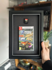 A framed display of Rocket League for Nintendo Switch, featuring the game case and disc mounted in a black shadow box with a white border. The frame is being held up against a gaming setup, with shelves displaying collectibles and gaming accessories in the background.