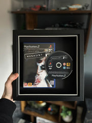 A framed display of Manuhunt for Playstation 2, featuring the game case and disc mounted in a black shadow box with a white border. The frame is being held up against a gaming setup, with shelves displaying collectibles and gaming accessories in the background.