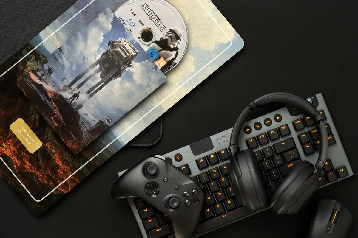 A styled flat-lay of a gaming setup on a dark surface, featuring a black mechanical keyboard with orange backlighting, black over-ear headphones, a black game controller, and a mouse. A large rectangular mouse pad with a Star Wars battle scene and a disc lies in the upper left.