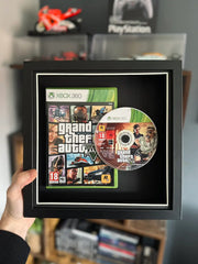 A framed display of Grand Theft Auto 5 for Xbox 360, featuring the game case and disc mounted in a black shadow box with a white border. The frame is being held up against a gaming setup, with shelves displaying collectibles and gaming accessories in the background.