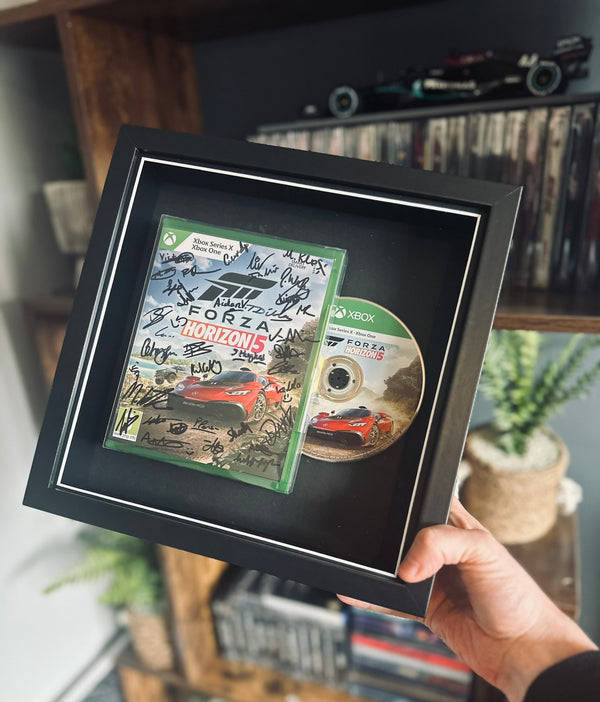 A framed display of Forza Horizon 5 for Xbox One, featuring the game case and disc mounted in a black shadow box with a white border. The game case is signed by a game development team. The frame is being held up against a gaming setup, with shelves displaying collectibles and gaming accessories in the background.