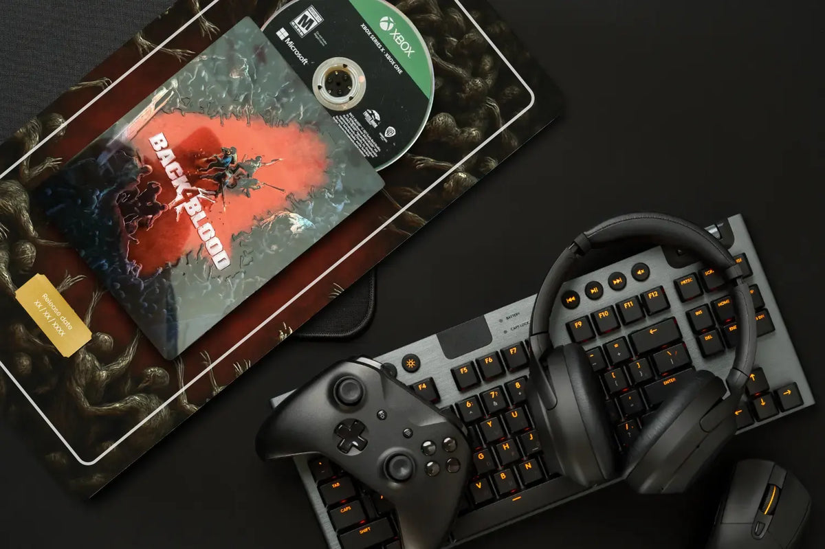 A gaming setup flat lay featuring a Black Back4Blood game disc, a game case, a wireless keyboard with orange backlighting, over-ear headphones, and a game controller on a dark surface.