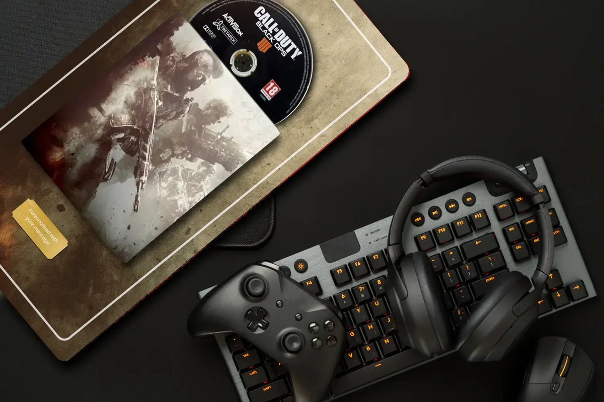 A flat-lay of a gaming setup on a dark surface, featuring a black and gray mechanical keyboard with orange backlighting, a black wireless controller, over-ear headphones, a mouse, and a Call of Duty game disc and box on a textured mat.