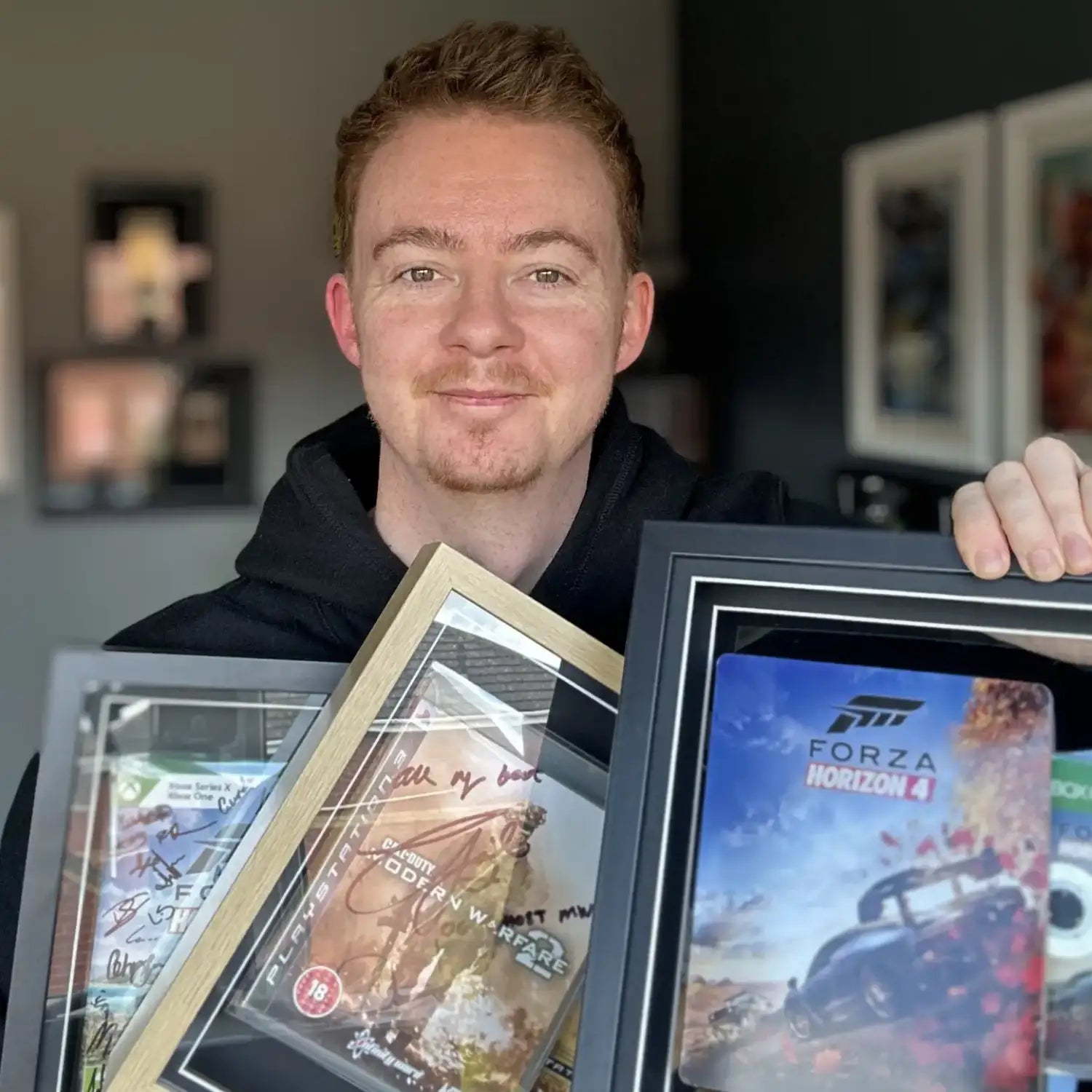 A man in a black hoodie holds up three framed memorabilia pieces, including a signed Forza Horizon 4 game and two autographed photo frames, in an indoor setting.