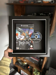 A framed display of TimeSplitters 2 for Nintendo GameCube, featuring the game case and disc mounted in a black shadow box with a white border. The frame is being held up against a gaming setup, with shelves displaying collectibles and gaming accessories in the background.