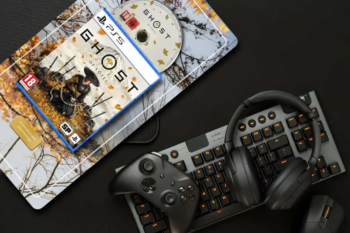 A PlayStation 5 Ghost Edition game case lies diagonally on a printed autumn-themed mat, with a PS5 disc beside it. The setup is styled on a dark surface with a black mechanical keyboard, black over-ear headphones, and a black game controller.