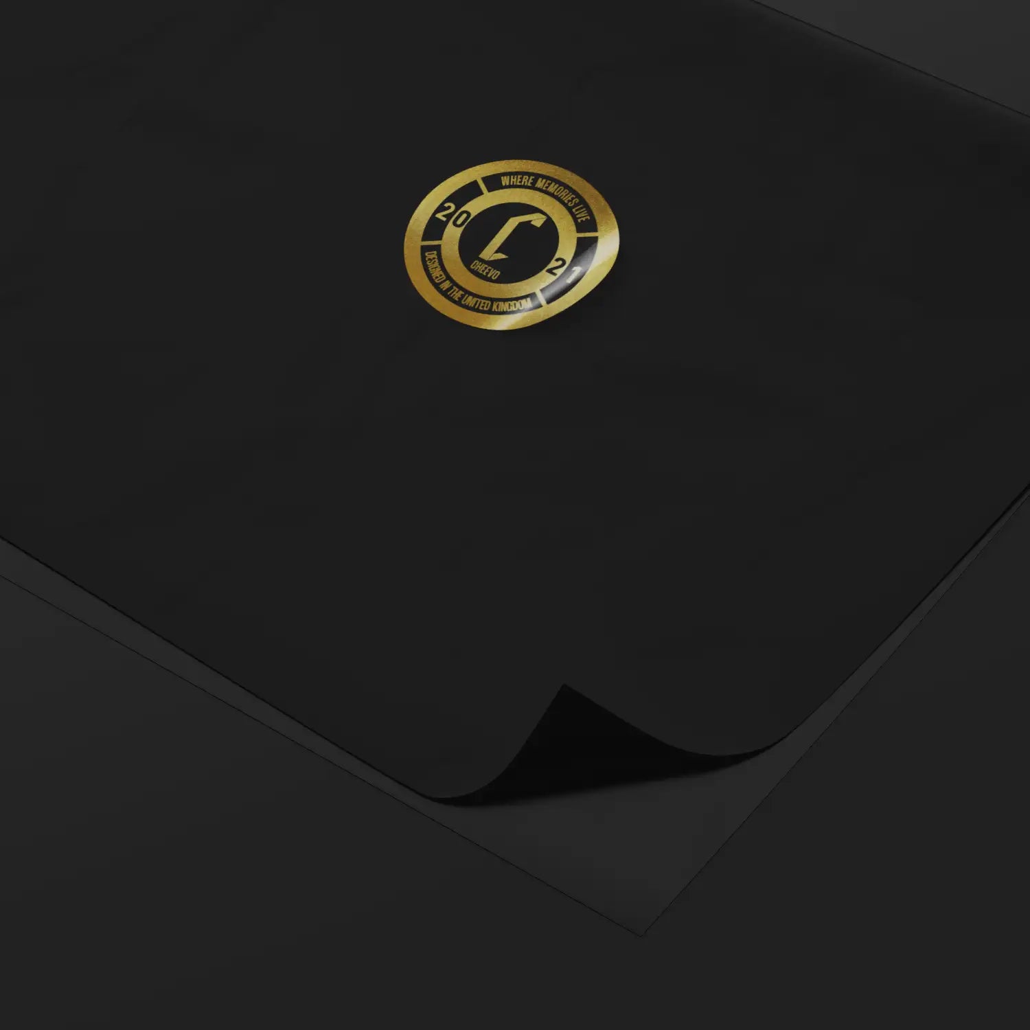 A close-up of a black rectangular product, likely a mat or pad, with a small circular gold logo centered near the top. The corner of the product is folded over, revealing a darker underside.