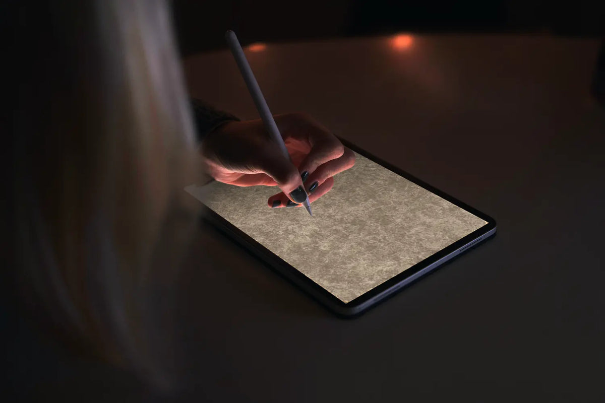 A close-up of a person's hand holding a stylus over a tablet screen, with soft warm lighting and blurred hair in the foreground on a dark tabletop.