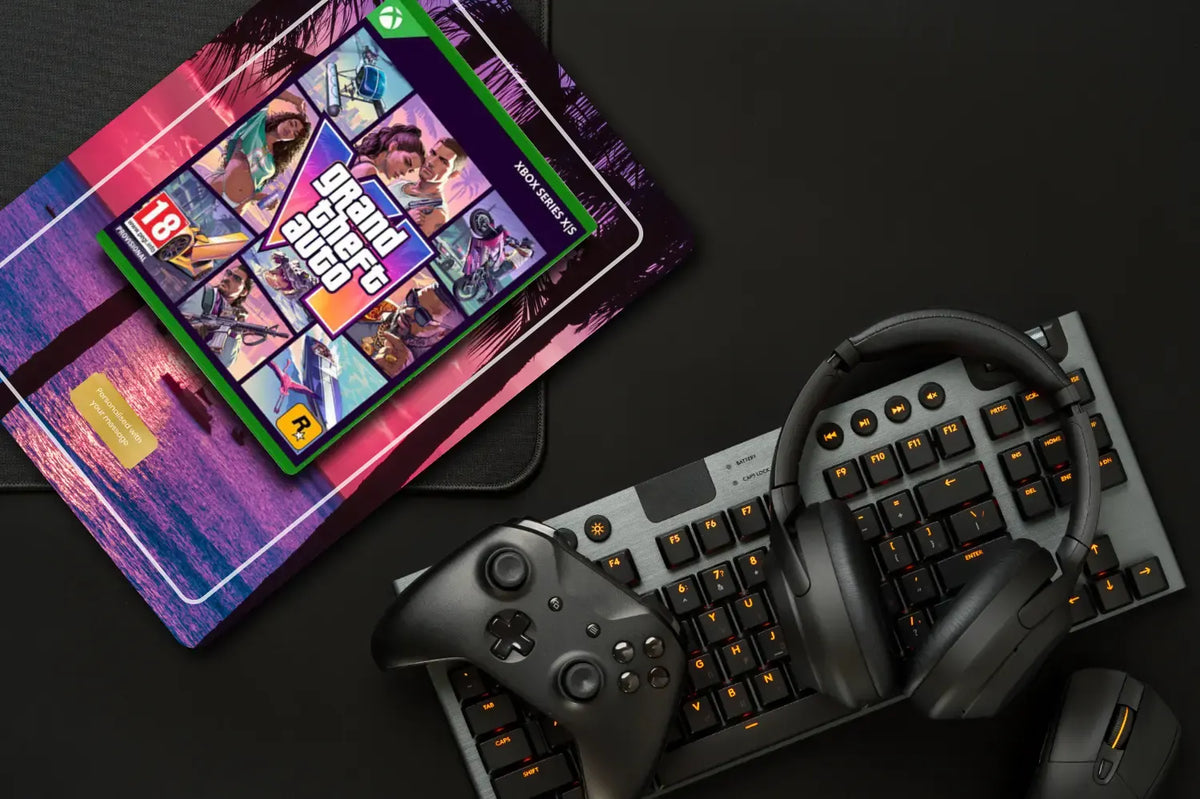 A gaming setup flat-lay featuring a black Xbox controller, a black mechanical keyboard with orange backlighting, over-ear headphones, and a mouse, alongside a printed magazine or booklet with a colourful Grand Theft Auto VI cover on a dark surface.