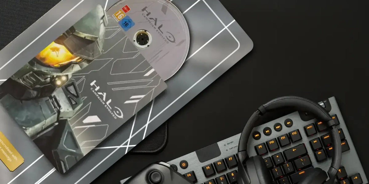 A Halo-themed desk setup featuring a printed card or insert with a Halo character illustration and disc, placed on a dark surface alongside a keyboard, headset, and controller. The Halo branding is visible on the disc and insert.