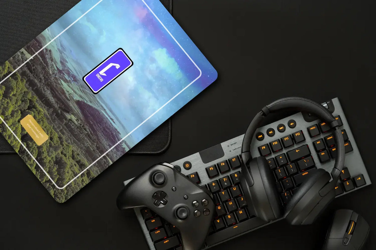 A top-down flat lay of gaming accessories on a dark surface: a black keyboard with orange backlighting, over-ear headphones, a game controller, and a mouse, alongside a card or case featuring a scenic print and a blue phone icon.