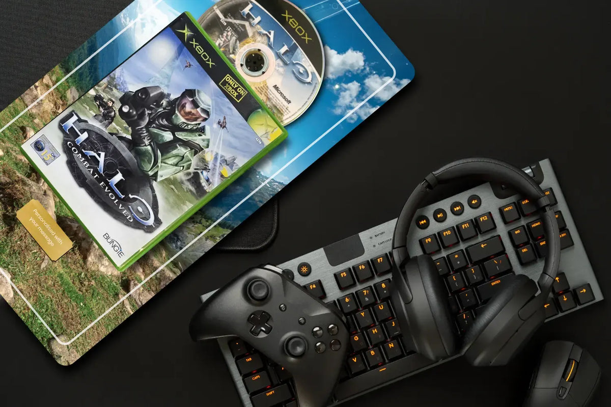 A gaming setup arranged on a dark surface, including a Halo game case, Halo disc, headset, keyboard, and controller. The Halo game packaging is the most prominent sold item, with Xbox branding visible on the case and disc.
