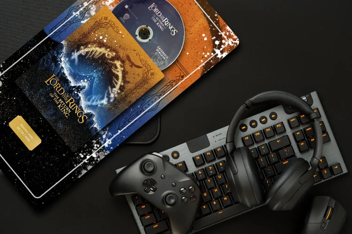 A styled flat-lay of gaming accessories on a dark surface: a black keyboard with orange backlighting, over-ear headphones, a black game controller, and a printed Lord of the Rings themed artwork or case featuring a disc. No person or real environment is present beyond the styled setup, so this fits Product/Packaging rather than Lifestyle.