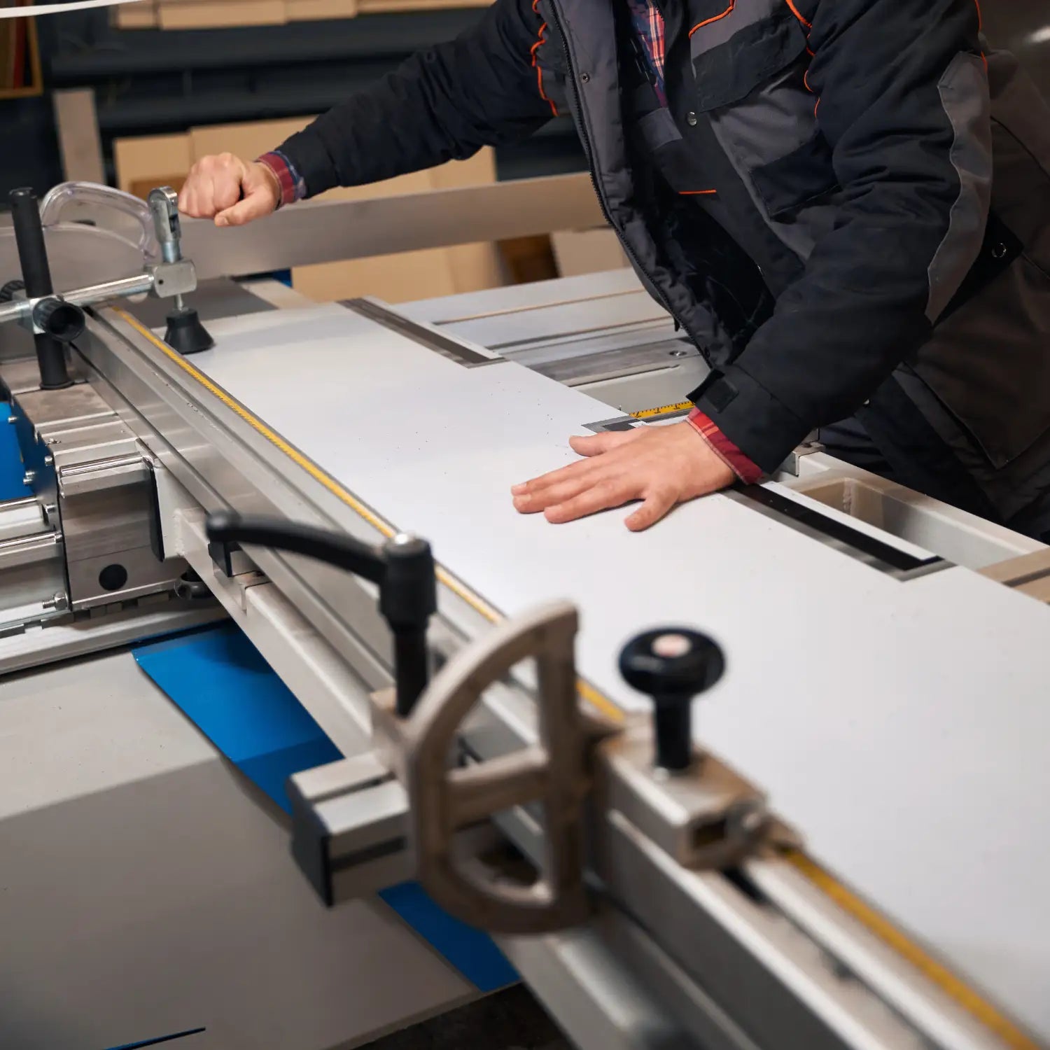 A person operating a large industrial table saw or cutting machine, guiding a white sheet of material with both hands. The machine has silver metal rails, black knobs, and a blue component underneath.