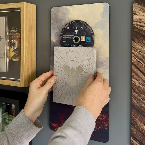 A person's hands are shown Destiny steelbook design off a decorative vertical display board featuring Destiny game art and a disc. To the left is a wooden framed display case.