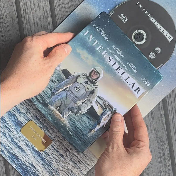 A person holds an Interstellar steelbook case with the Blu-ray disc partially visible behind it, photographed from above on a gray wood surface. The cover shows an astronaut standing in water, with the Interstellar title and cast names visible.