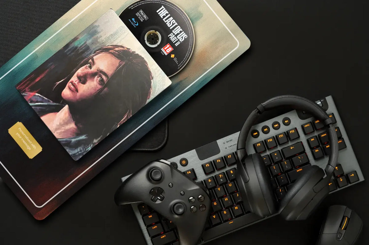 A gaming setup flat-lay featuring a black mechanical keyboard with orange backlighting, black over-ear headphones, a black game controller, and a custom printed artwork showcasing artwork from The Last of Us with a disc on top, all arranged on a dark surface.