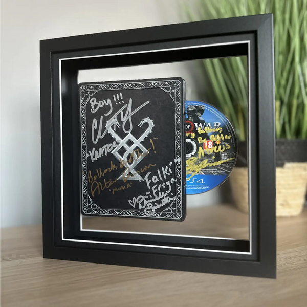 A black shadow box frame displayed on a wooden surface, containing a black chalkboard-style sign with white and gold signatures and a blue PlayStation 4 disc tucked behind it.