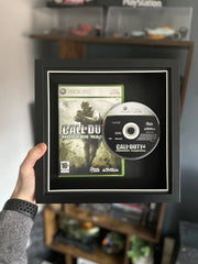 A framed display of Call of Duty 4: Modern Warfare for Xbox 360, featuring the game case and disc mounted in a sleek black shadow box with a white border. The frame is being held up in front of a gaming setup with shelves displaying collectibles and gaming accessories in the background.